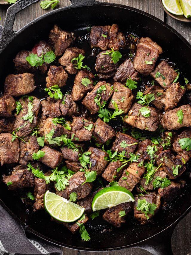Overhead view of Hatch chile steak bites garnished with cilantro and lime wedges in a cast-iron skillet.