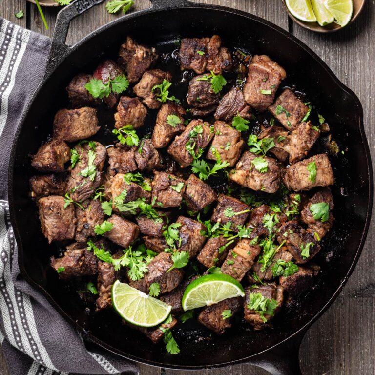 Overhead view of Hatch chile steak bites garnished with cilantro and lime wedges in a cast-iron skillet.