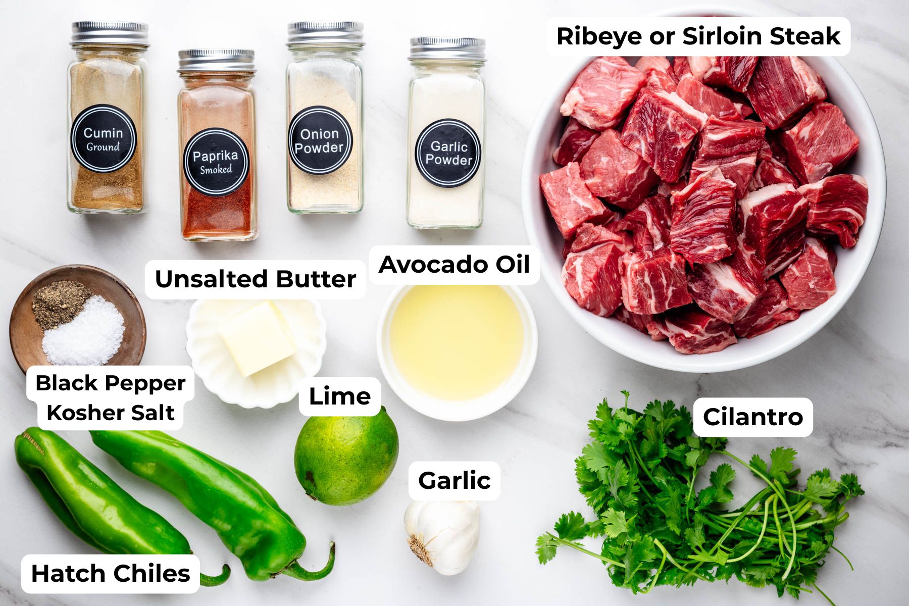 Labeled overhead view of ingredients for Hatch chile steak bites, including cubed steak, Hatch chiles, butter, avocado oil, lime, garlic, cilantro, and seasonings.