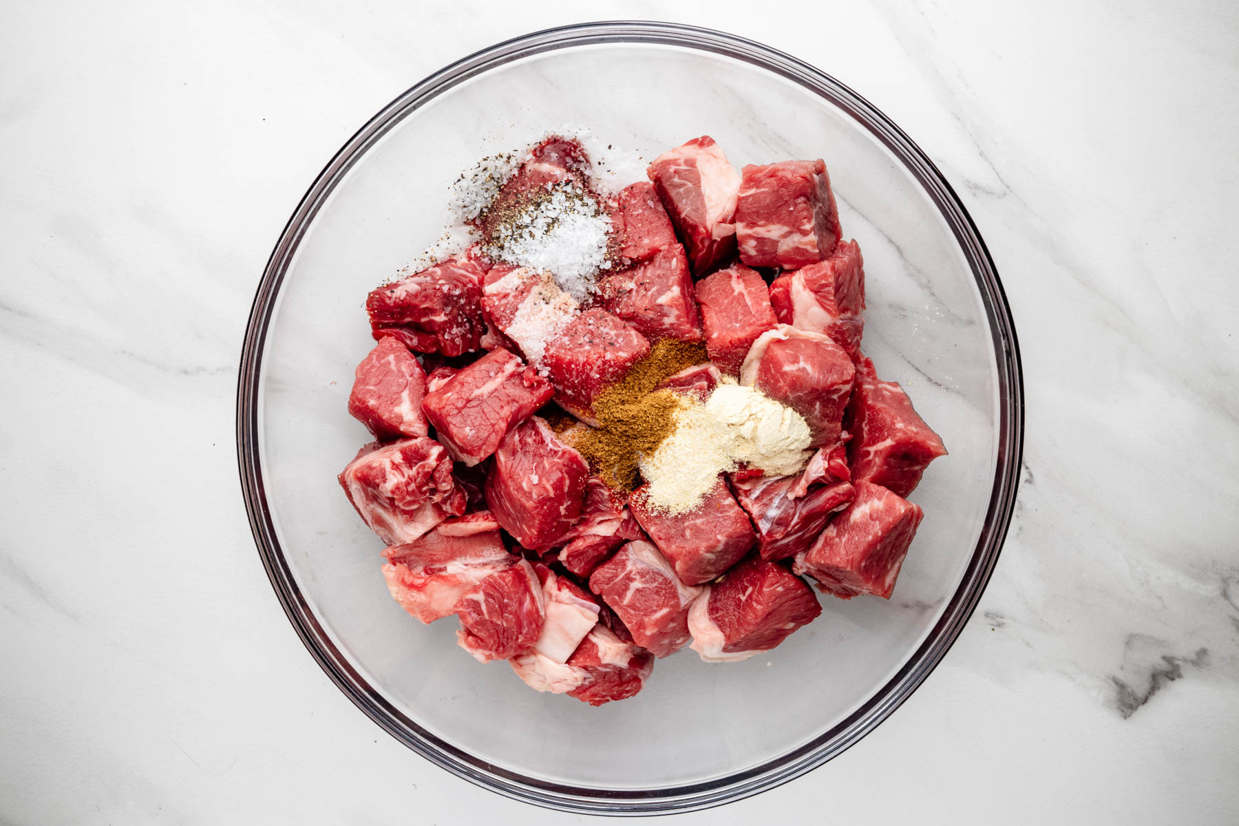Cubed ribeye steak topped with kosher salt, black pepper, cumin, smoked paprika, garlic powder, and onion powder in a glass bowl.