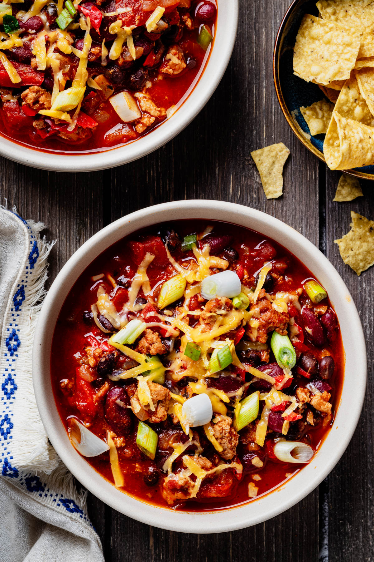 Overhead view of healthy turkey chili topped with shredded cheese and green onions, served with tortilla chips.