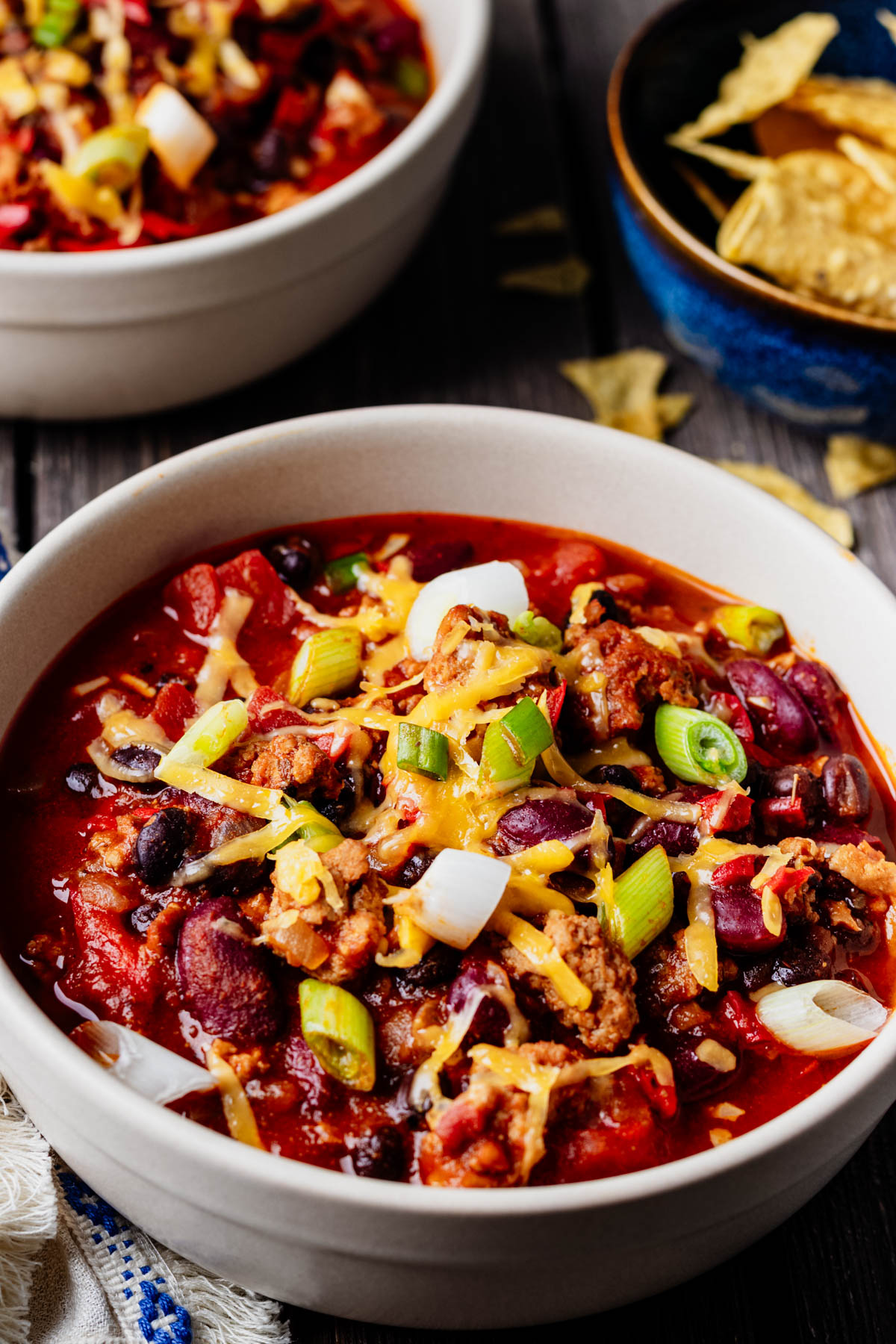 Close-up of healthy turkey chili topped with shredded cheese and green onions, with tortilla chips in the background.