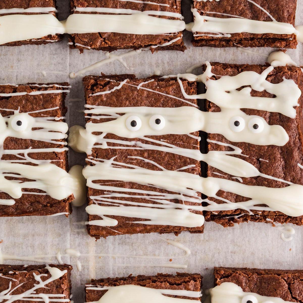 Close-up of mummy brownies decorated with white chocolate drizzle and candy eyes on parchment paper.