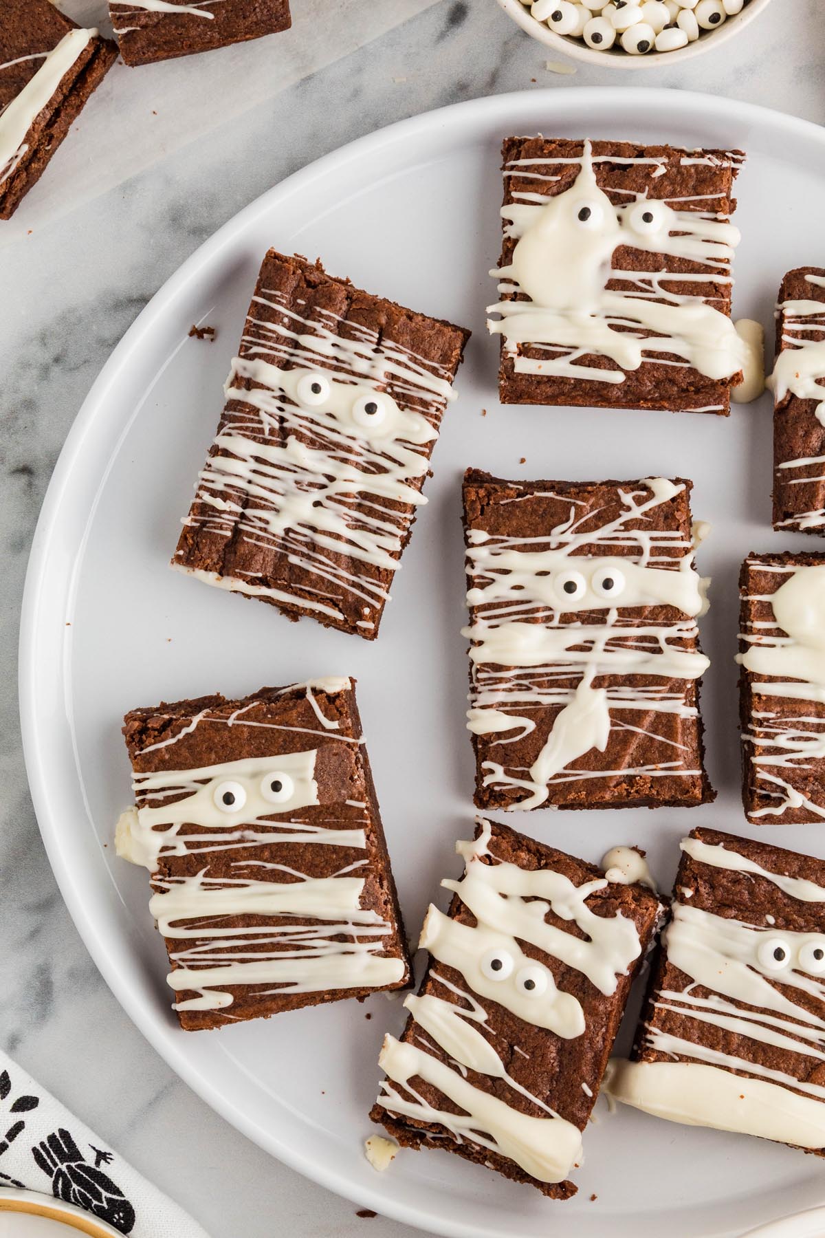 Mummy brownies arranged on a white serving platter, decorated with white chocolate drizzle and candy eyes.