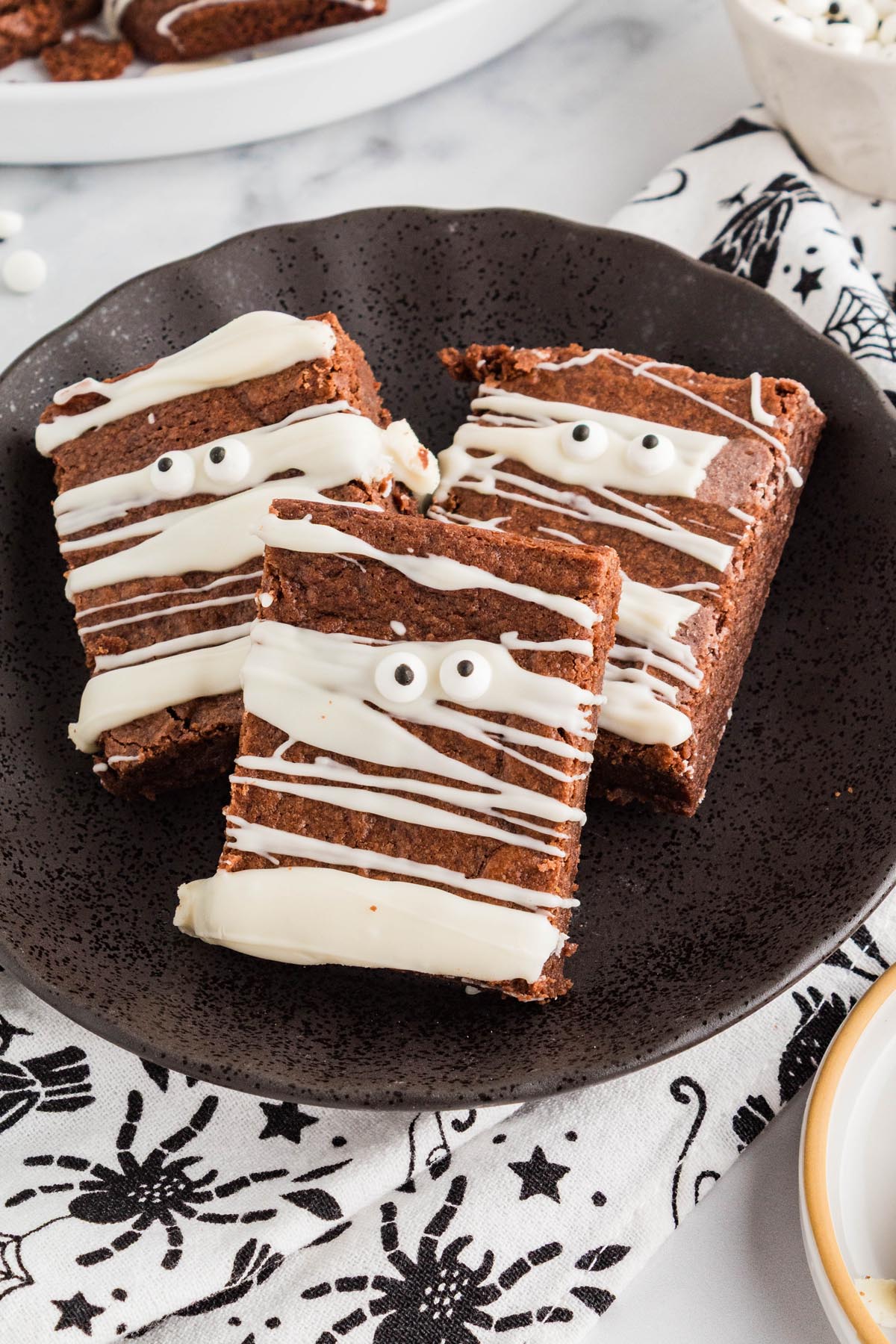 Three mummy brownies served on a black plate with white chocolate drizzle, candy eyes, and a Halloween-patterned napkin.