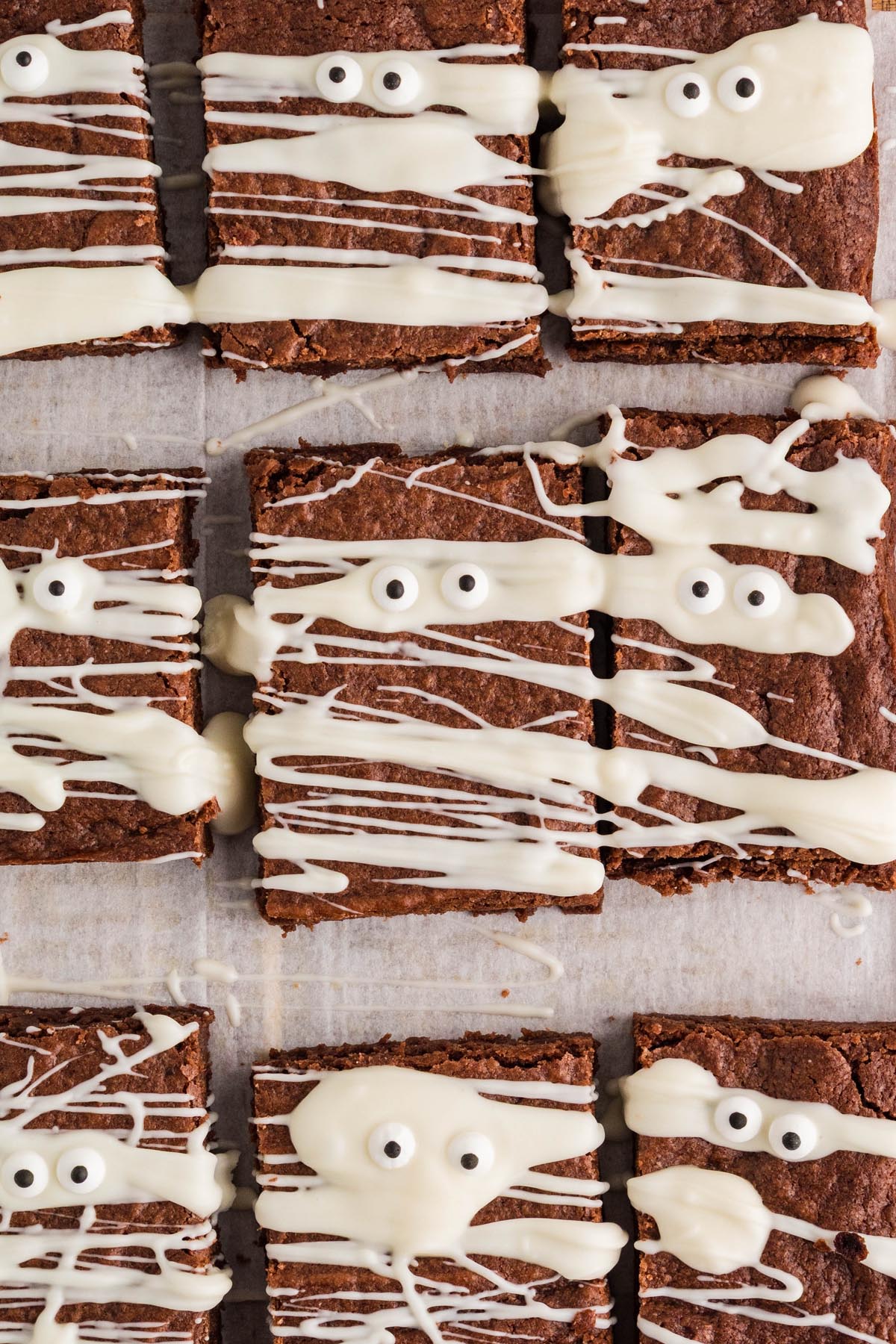 Mummy brownies arranged on parchment paper, each decorated with white chocolate drizzle and candy eyes.