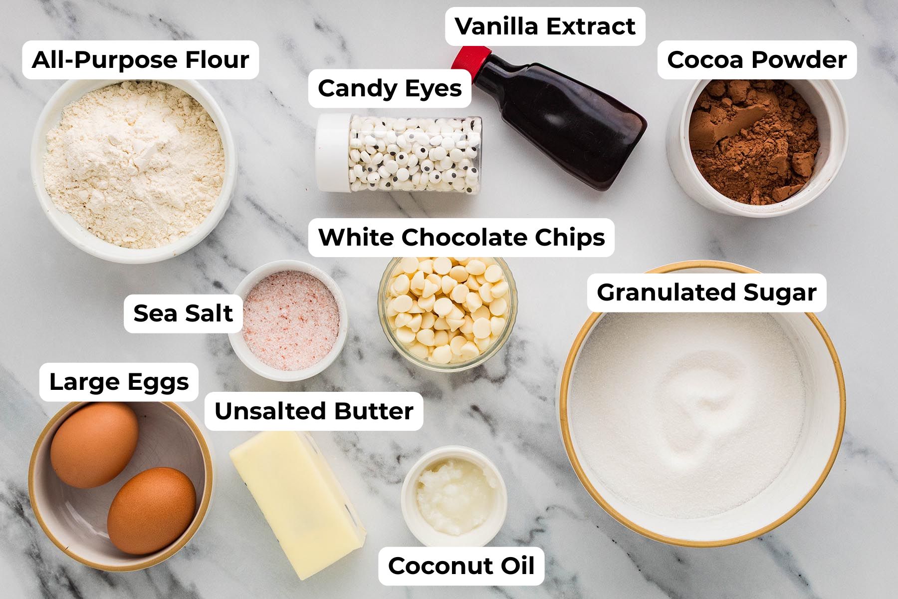 Labeled ingredients for mummy brownies, including all-purpose flour, candy eyes, vanilla extract, cocoa powder, sea salt, white chocolate chips, granulated sugar, eggs, unsalted butter, and coconut oil.