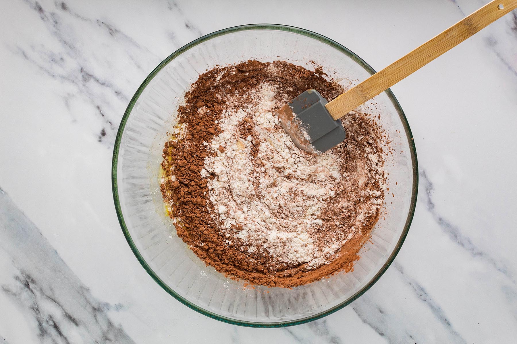 Flour, cocoa powder, and salt added to the wet brownie ingredients in a glass bowl.