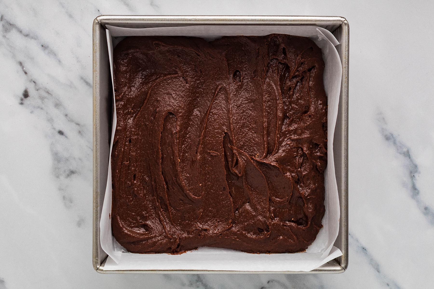 Chocolate brownie batter spread in a parchment-lined square metal baking pan.