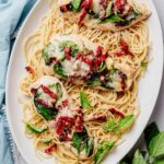 Overhead view of sheet pan Tuscan chicken topped with spinach, sun-dried tomatoes, melted Parmesan, and fresh basil, served over spaghetti on a white platter.