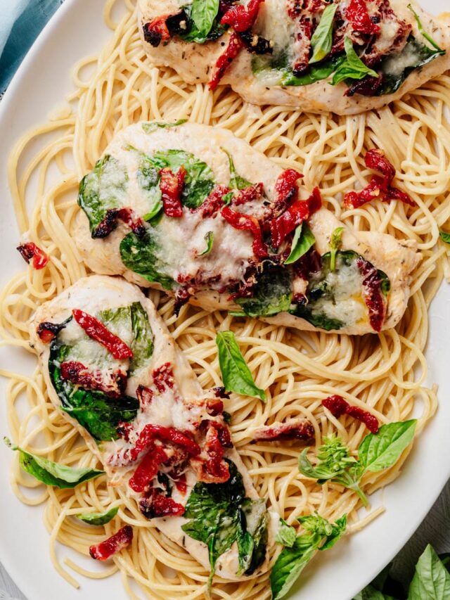 Overhead view of sheet pan Tuscan chicken topped with spinach, sun-dried tomatoes, melted Parmesan, and fresh basil, served over spaghetti on a white platter.