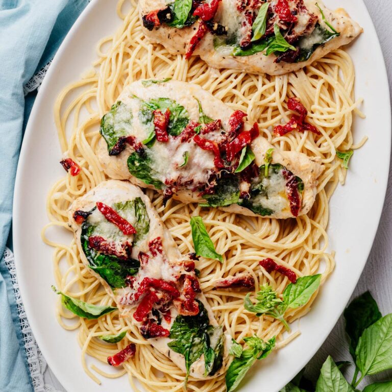Overhead view of sheet pan Tuscan chicken topped with spinach, sun-dried tomatoes, melted Parmesan, and fresh basil, served over spaghetti on a white platter.