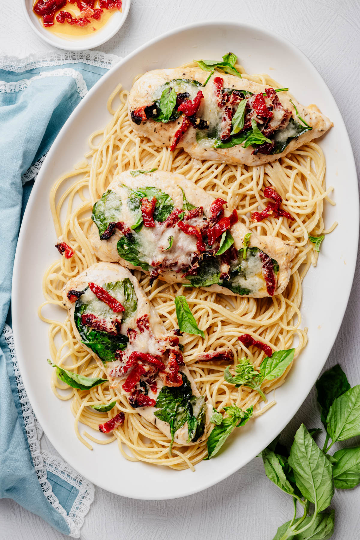 Overhead view of three Tuscan chicken cutlets with spinach, sun-dried tomatoes, melted Parmesan, and fresh basil, served over spaghetti on a white platter.