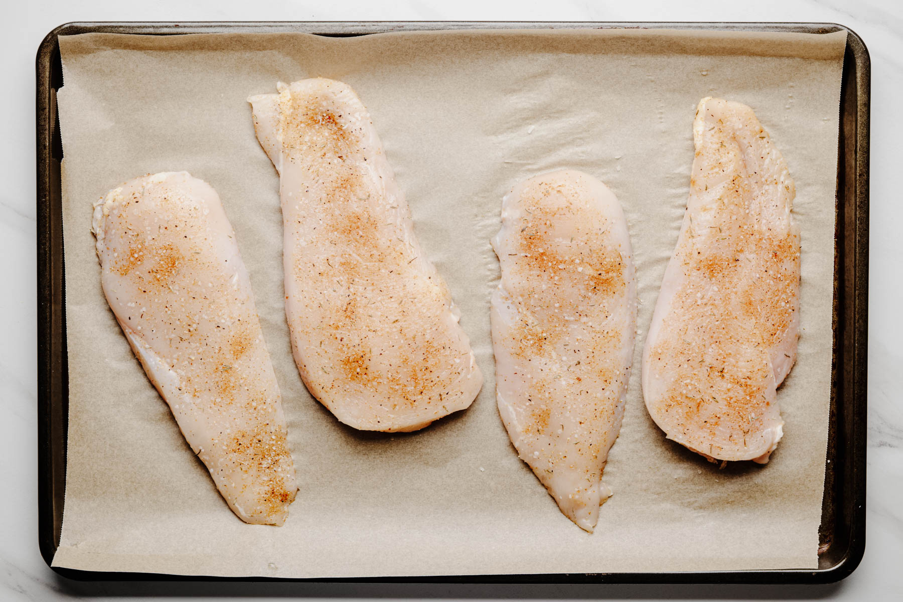 Four raw chicken cutlets lightly seasoned with garlic herb dry brine on a parchment-lined sheet pan.