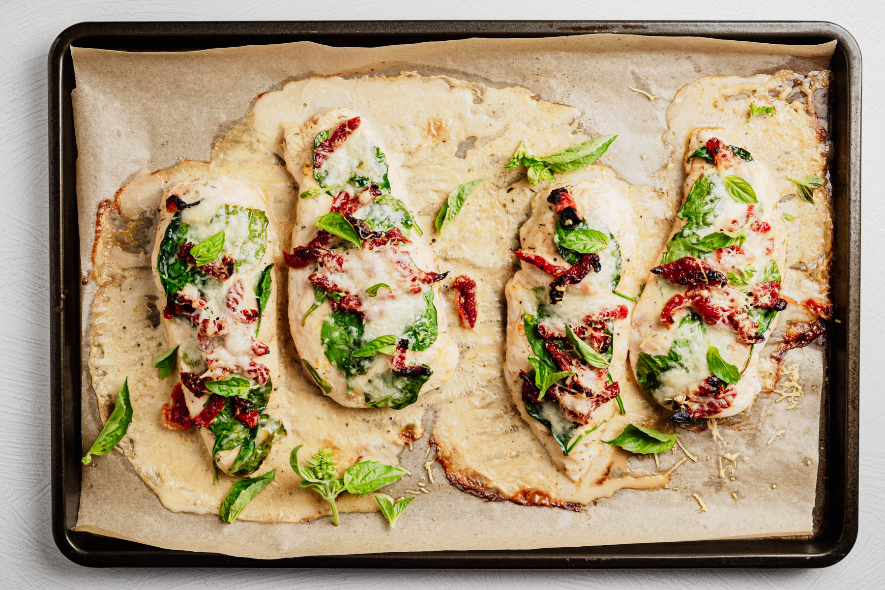 Tuscan chicken cutlets topped with spinach, sun-dried tomatoes, melted Parmesan, Alfredo sauce, and fresh basil on a sheet pan.
