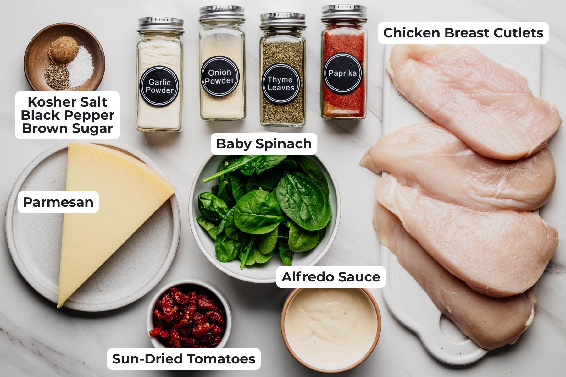 Overhead view of the ingredients for sheet pan Tuscan chicken, including chicken breast cutlets, Alfredo sauce, baby spinach, sun-dried tomatoes, Parmesan, and seasonings, with each ingredient labeled.